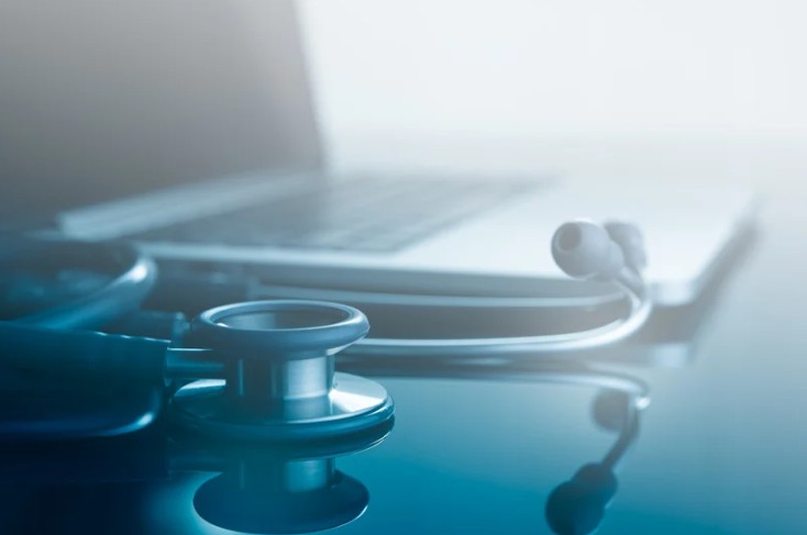 Estetoscopio apoyado sobre una superficie reflectante junto a un ordenador portátil, simbolizando la conexión entre tecnología y atención médica